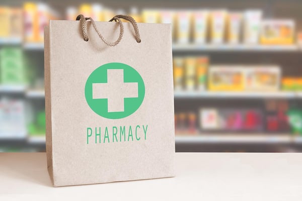 Etualalla paperikassi, jossa teksti Pharmacy ja vihreällä taustalla oleva ruskea risti, taustalla hyllyillä voiteita ja lääkkeitä.